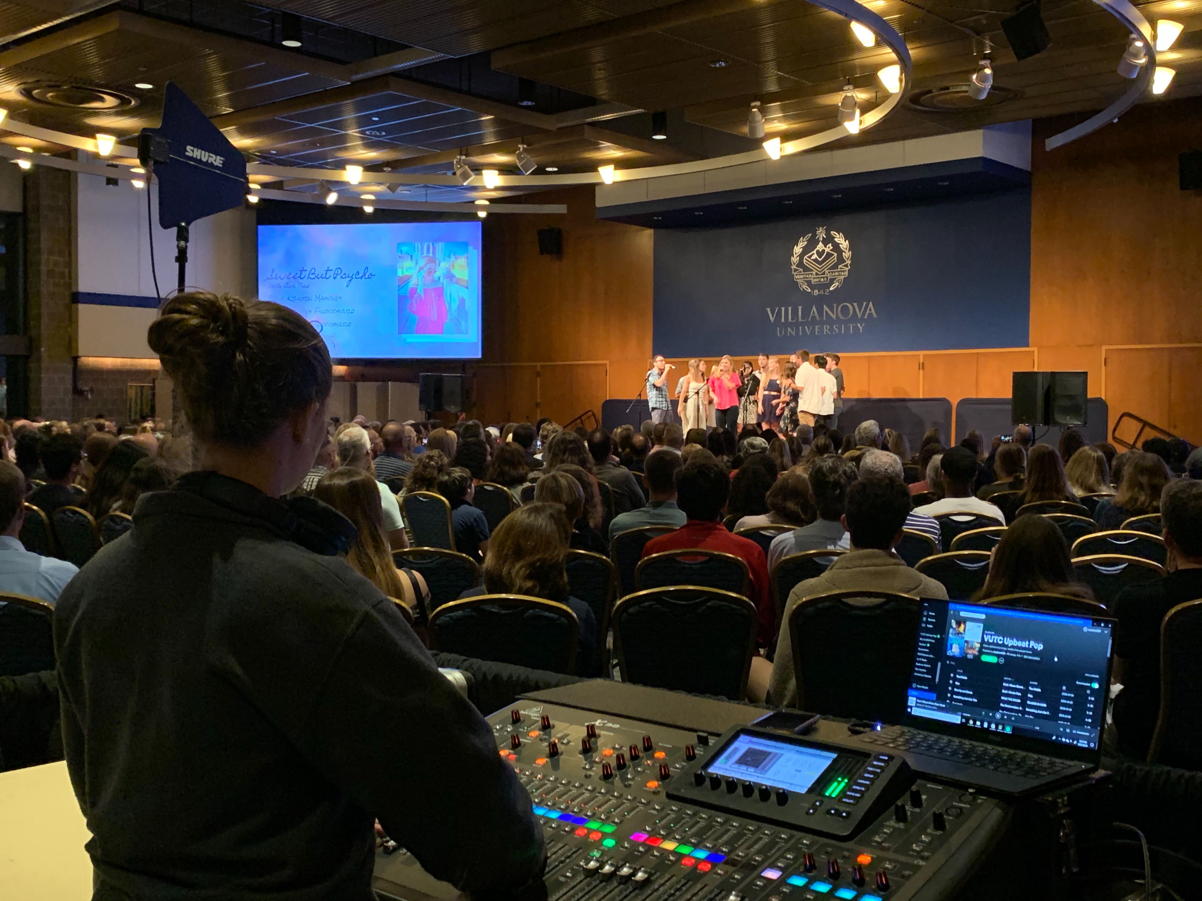 Anna Schmult mixing an a cappella concert. She stands at the back of a room full of people, watching the singers on stage.